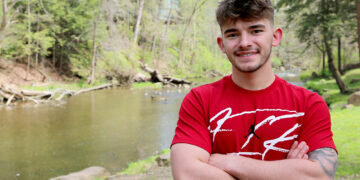 male student standing with arms crossed in front of creek