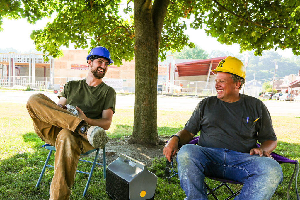 two men in yellow hard hats sitting outside eating lunch at a construction site