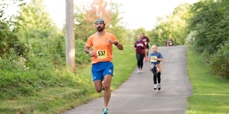 man with beard running in outdoor race wearing orange shirt, blue shorts, backwards teal cap with the number 537 on his race bib
