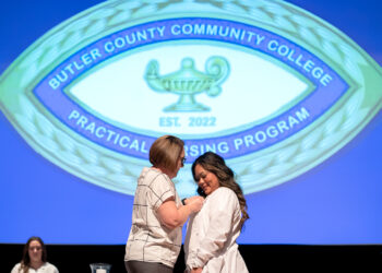 two women on stage at nurses pinning ceremony. woman on right wearing white nurses uniform recieves pin from the woman on the left. graphic projected in background reads "butler county community college practical nursing program EST. 2022"