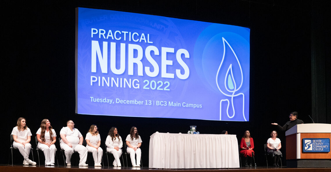 group of nurses in white scrubs on stage at their pinning ceremony, with speaker at podium and screen behind projects an image with a graphic candle with text that reads "Practical Nurses Pinning 2022. Tuesday, December 13 | BC3 Main Campus"
