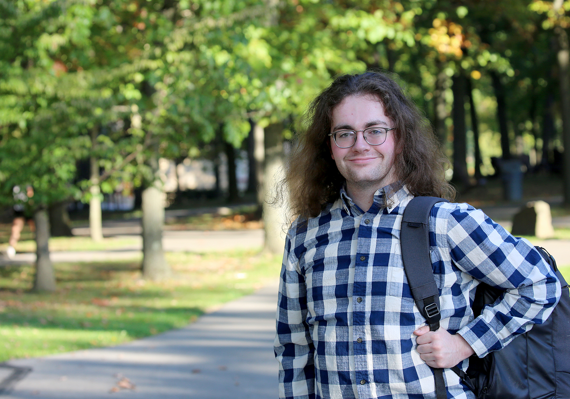 portrait of a man with long hair and glasses with a plaid shirt and backpack standing outside a path lined with oak trees